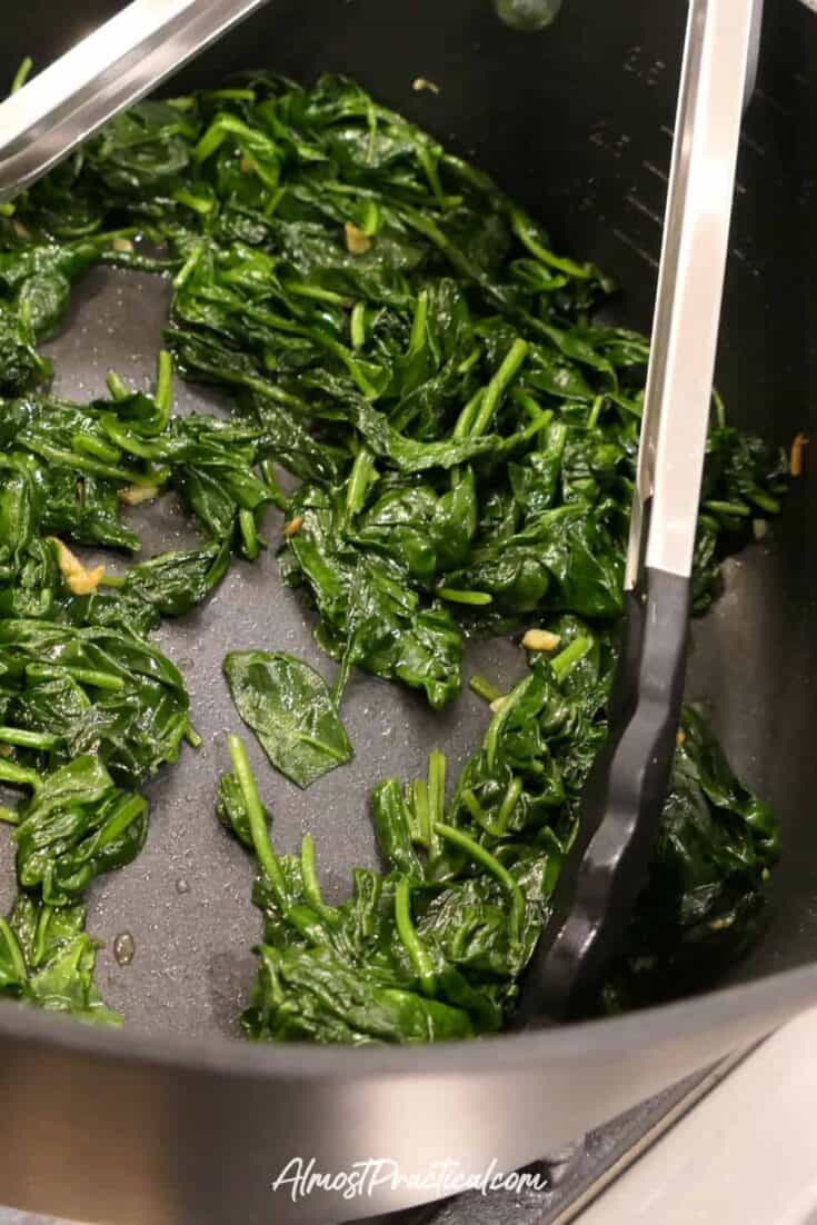 Photo showing wilted spinach with garlic in a nonstick pan with long handled silicone tipped tongs.